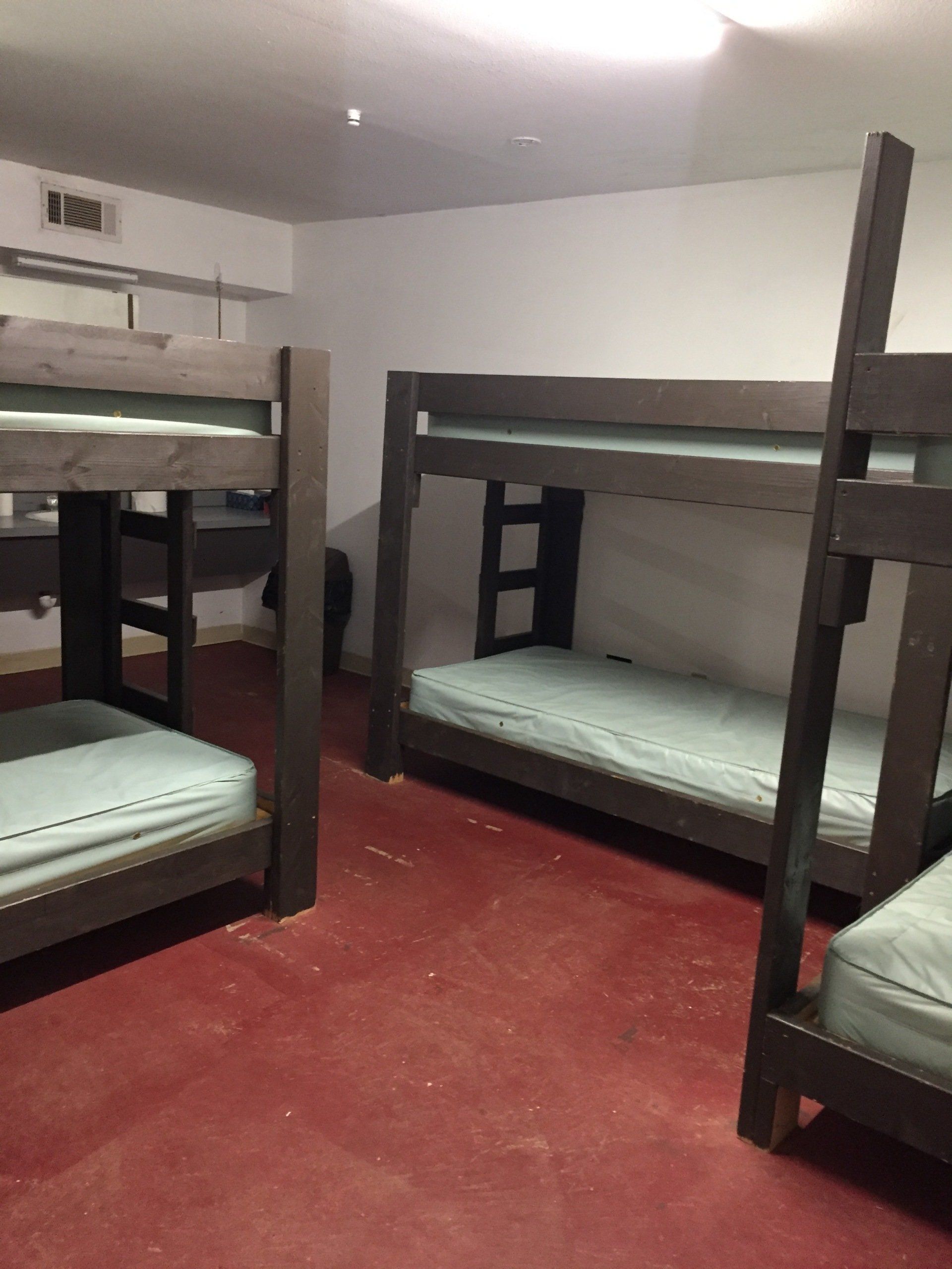 Four wooden bunk beds in a room with red floor, white walls, and a ceiling vent.