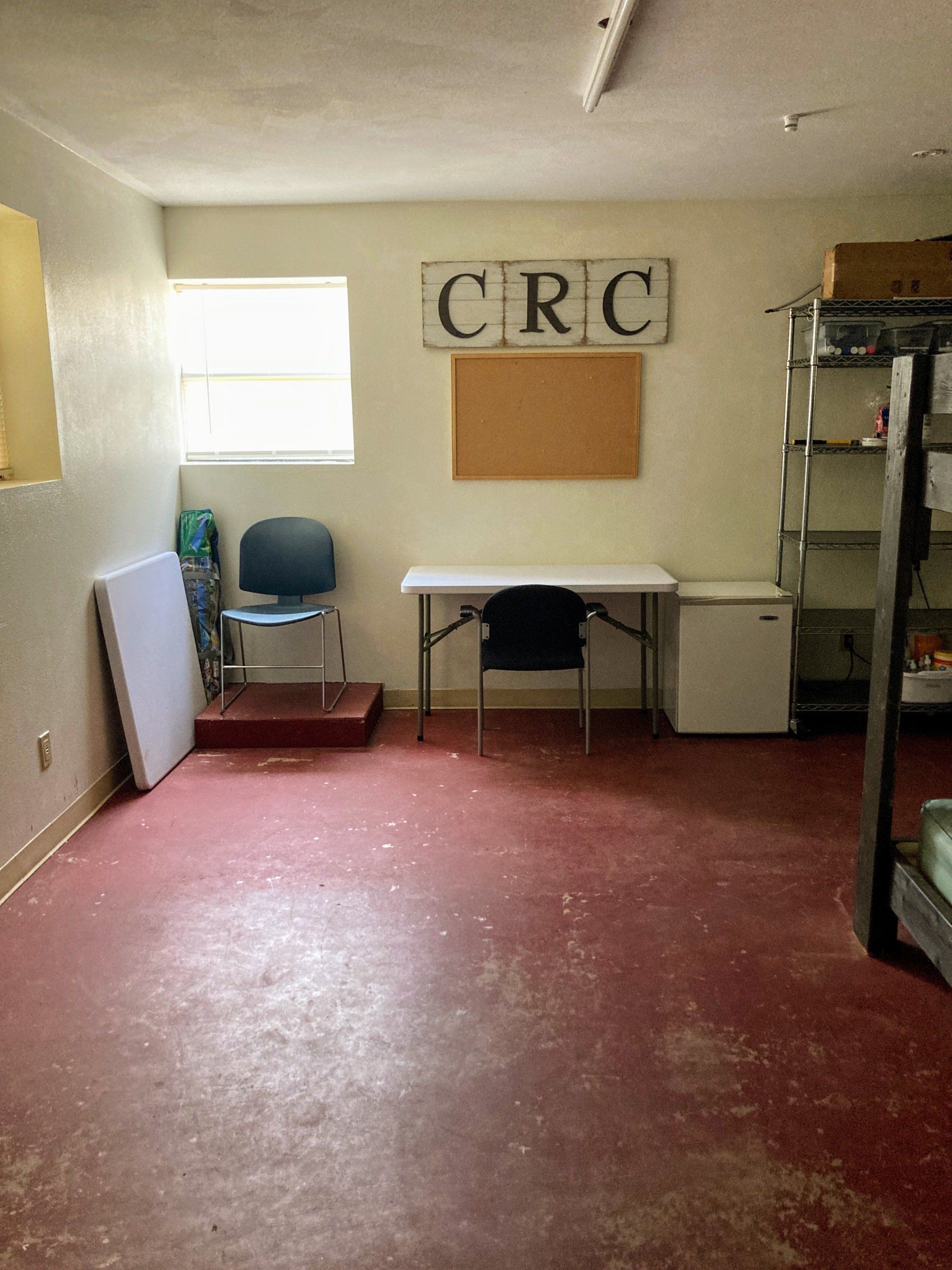 An empty room with red floor, white walls, table, chair, corkboard, and a shelf. 