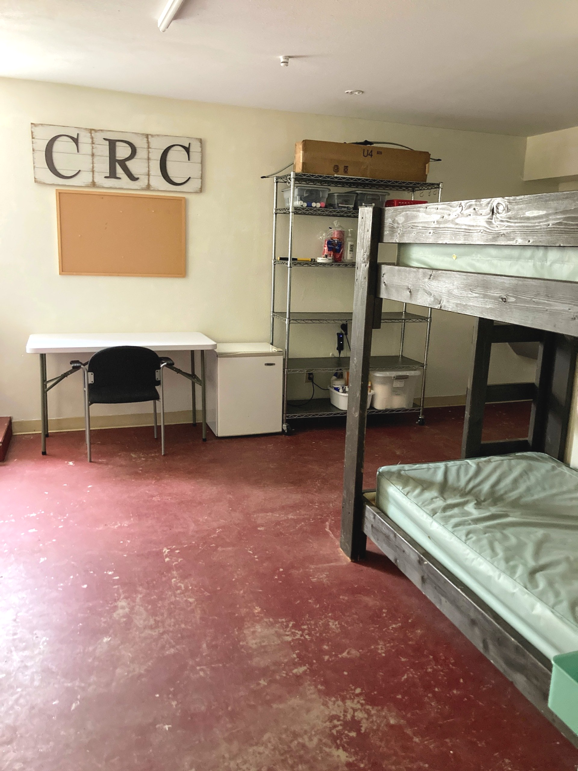 A dorm room with red floor, bunk beds, desk, mini-fridge, and storage shelves. 