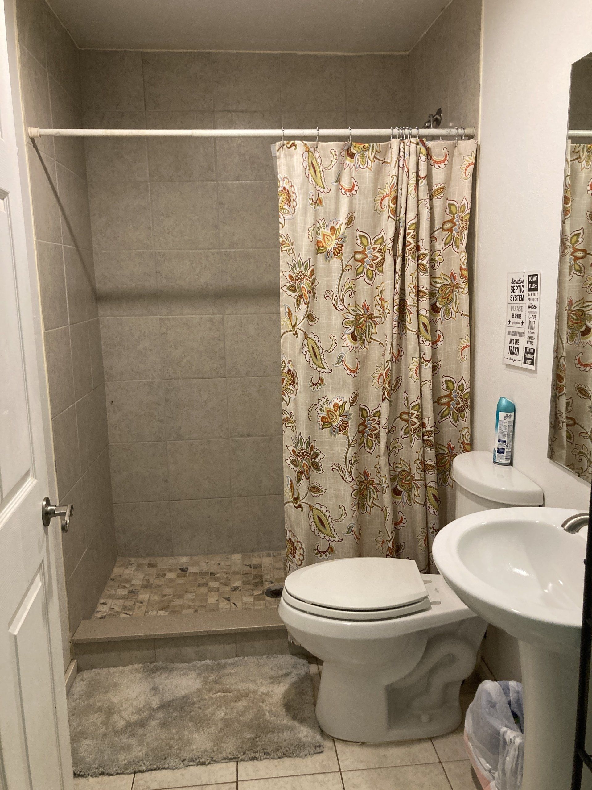 Bathroom with a shower, toilet, and sink. Beige shower curtain with floral pattern. Light gray tiles.