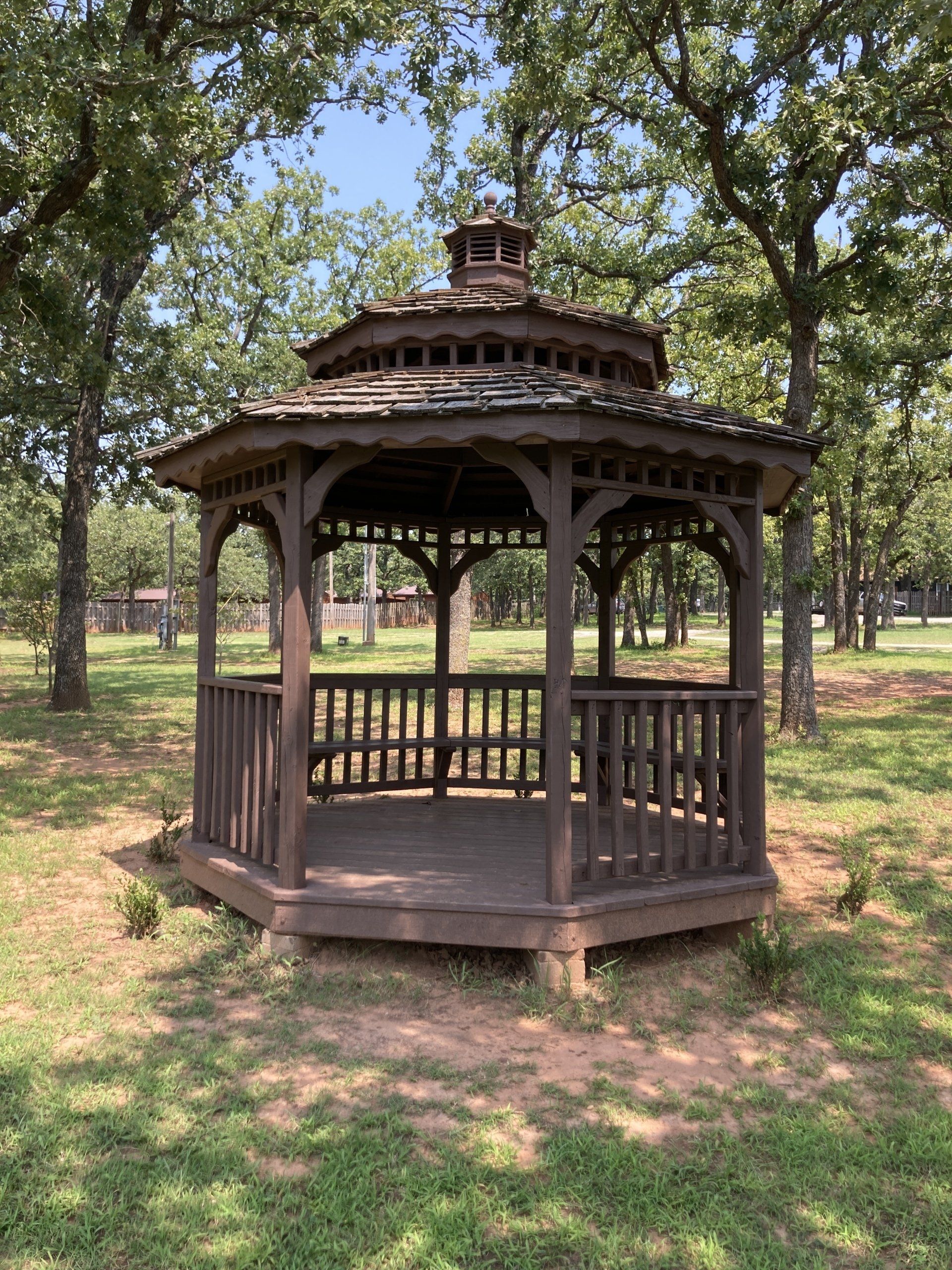 Wooden gazebo in a park, surrounded by trees and grass, under a blue sky.