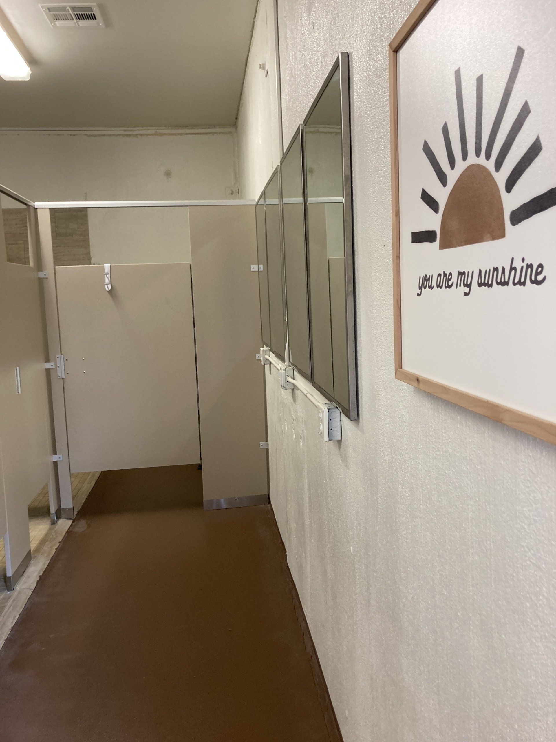 A public restroom with stalls on the left, mirrors, and a decorative sun-themed print on the wall.