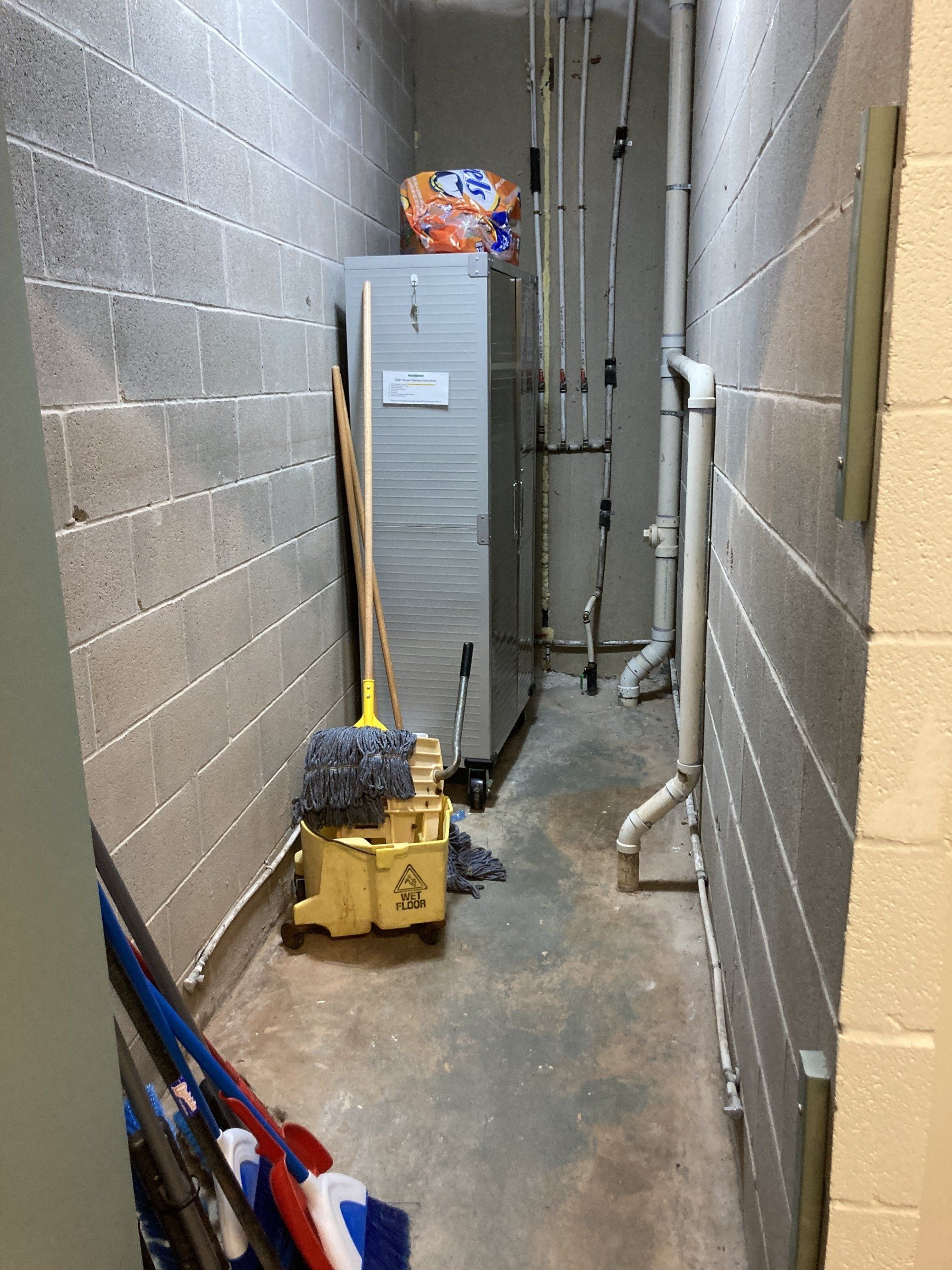 Narrow utility hallway with gray cinder block walls, pipes, and cleaning supplies.