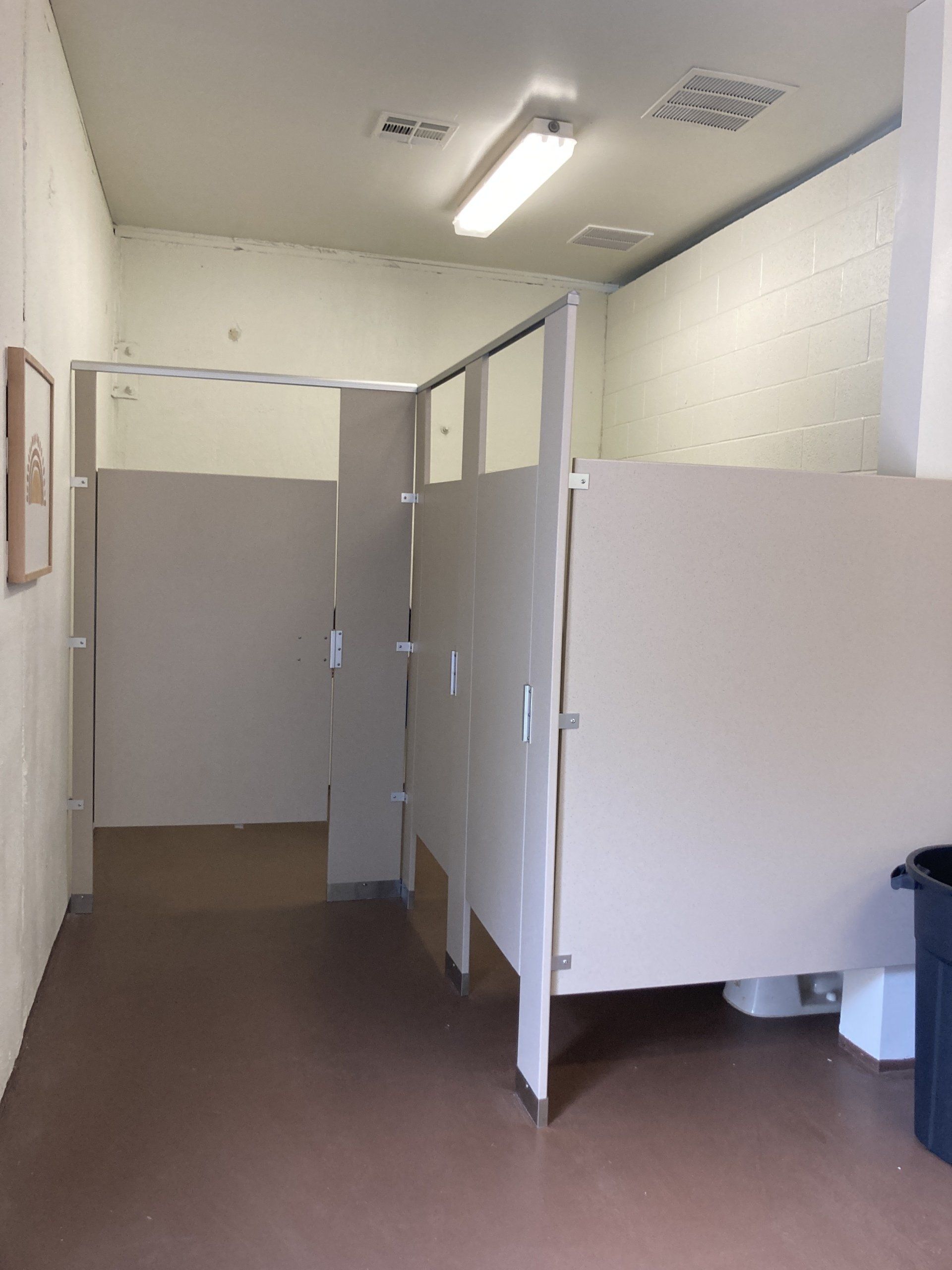 Public restroom with three stalls, light tan walls, and brown floor.