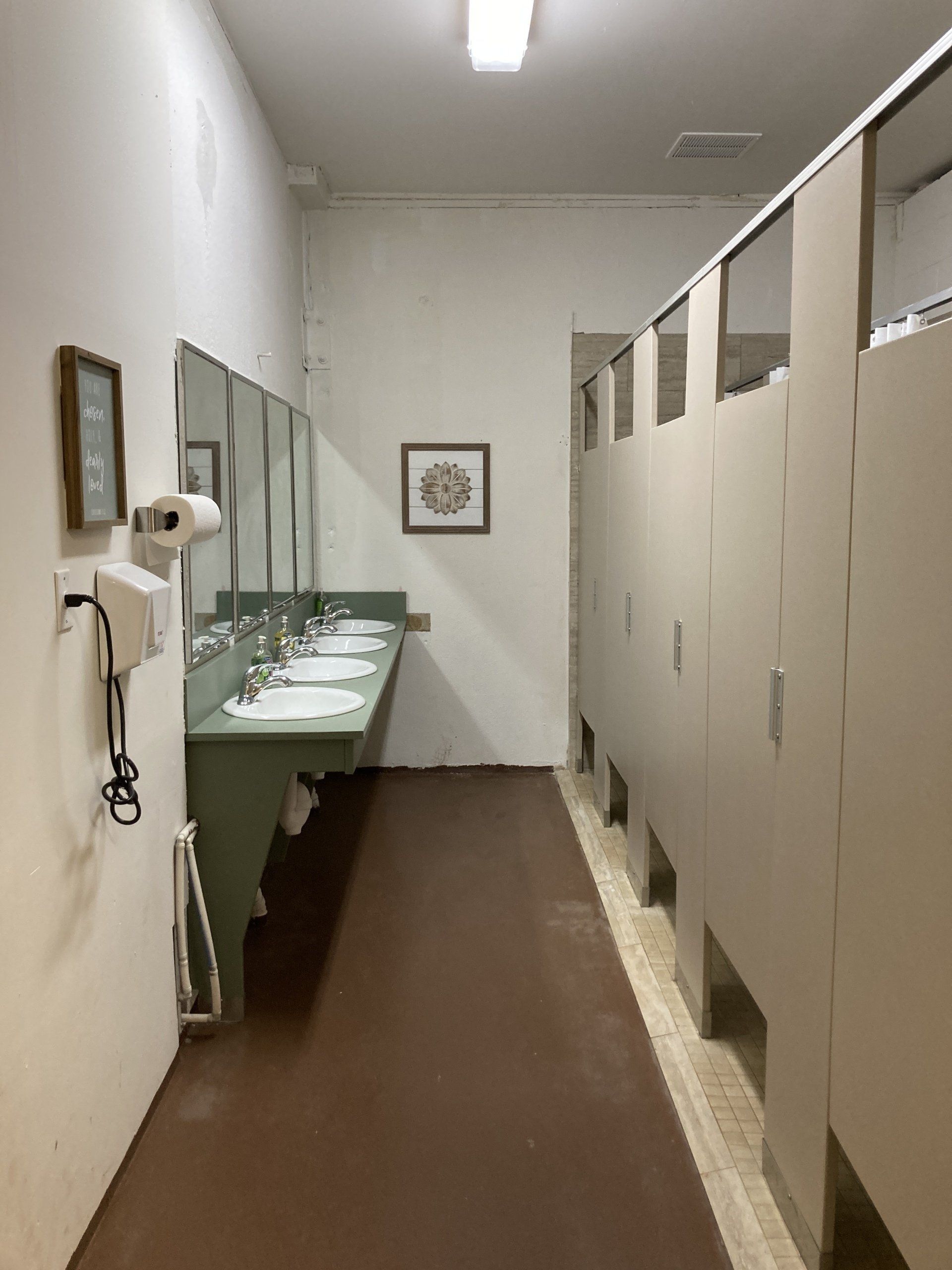 Bathroom with a row of sinks, mirrors, and stalls. The floor is brown and the walls are light.