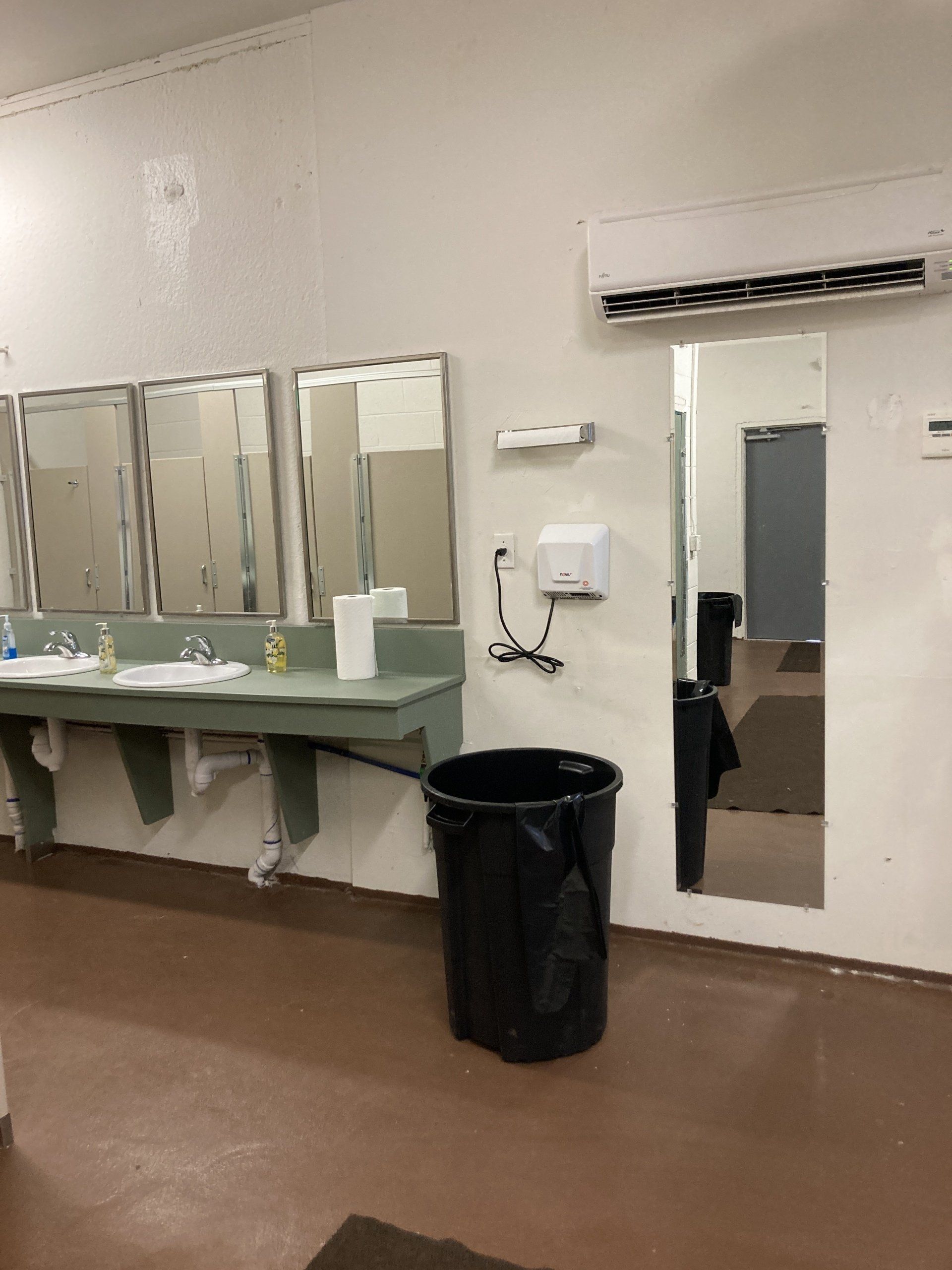 Public restroom with sinks, mirrors, and a trash can. White walls, brown floor.