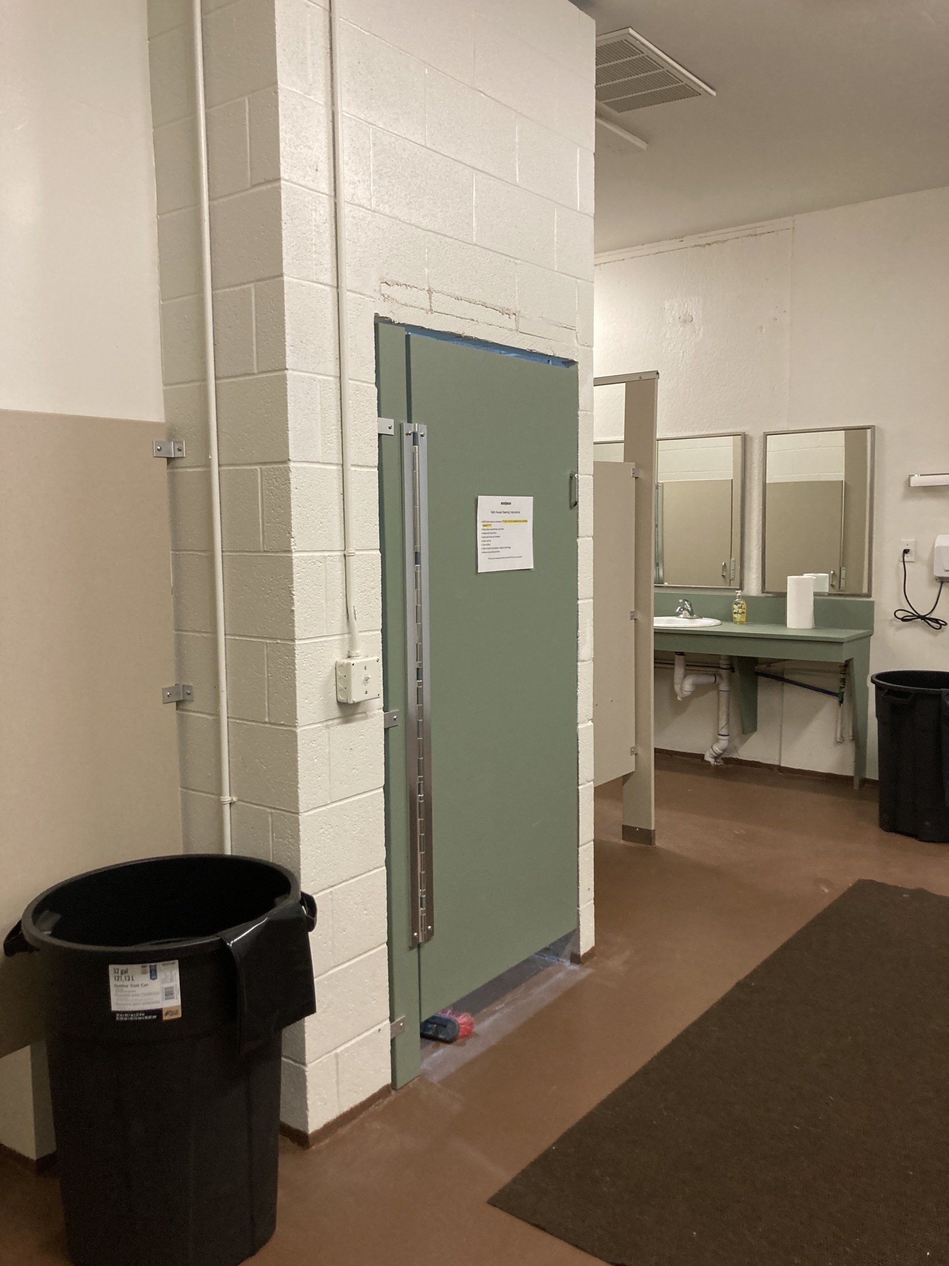 Public restroom entrance; green door, trash can, sinks, white brick walls.