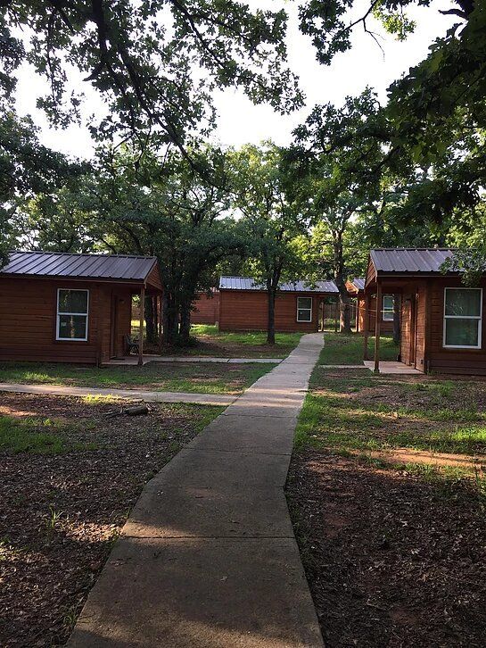 Concrete path leads between cabins, under trees. Brown cabins with metal roofs in a wooded area.