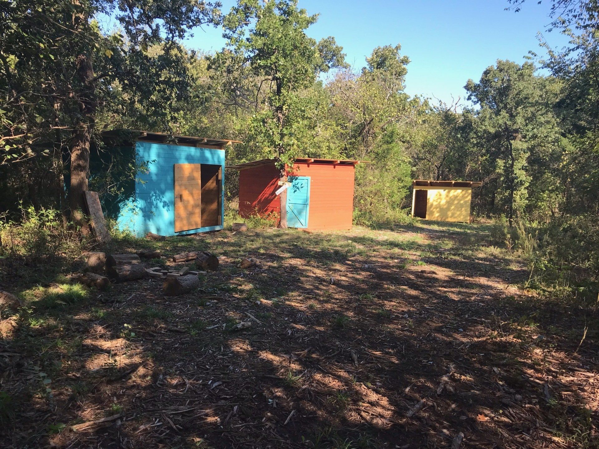 Three colorful small structures in a wooded area; blue, red, and gold, each with open doorways.
