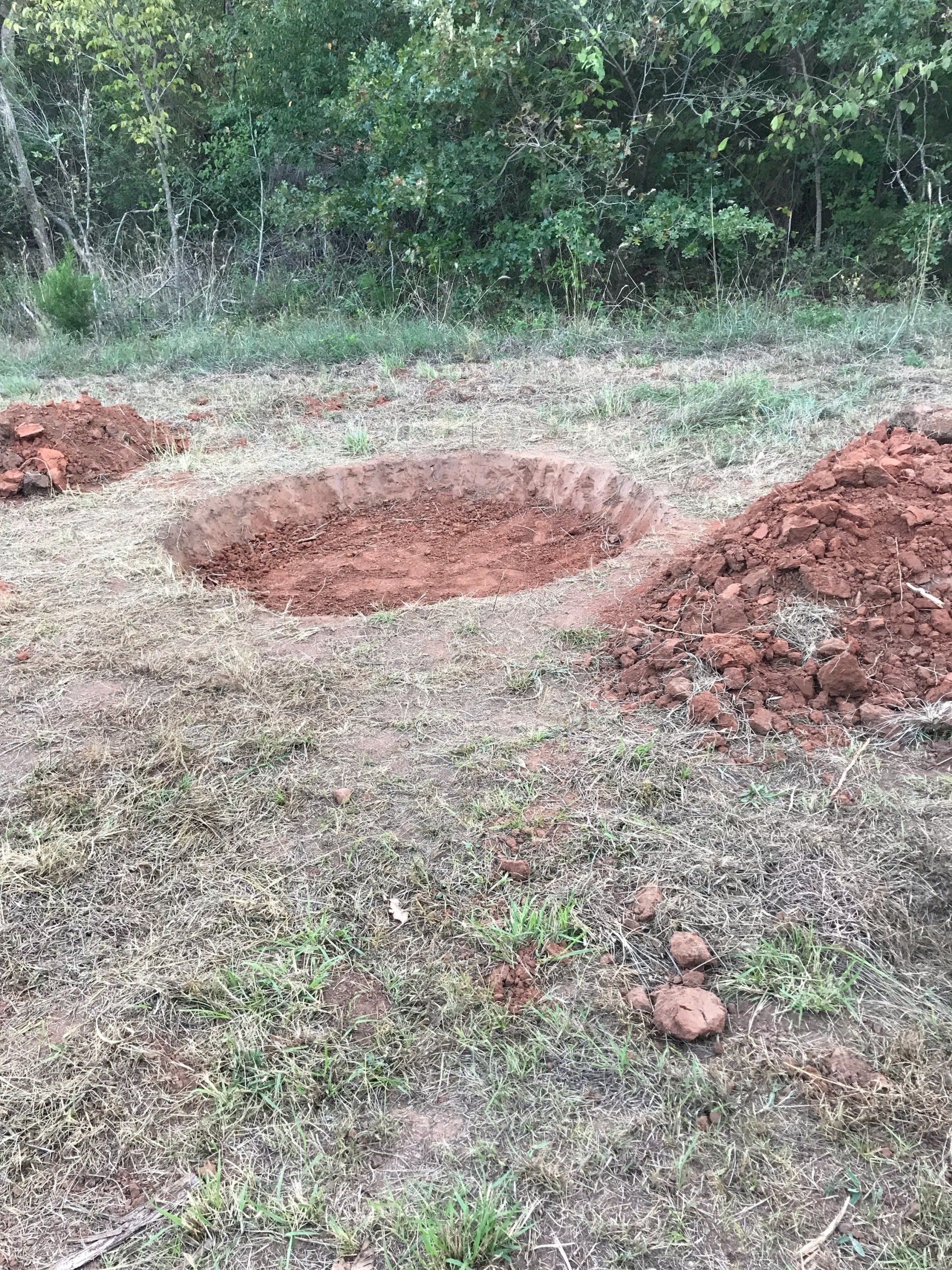 Holes dug in grassy ground with piles of reddish soil nearby, trees in the background.