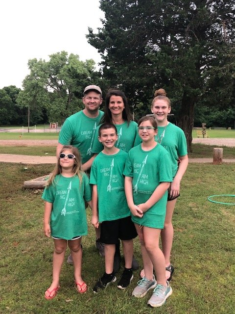 Family wearing green t-shirts outdoors; smiling, near a tree.