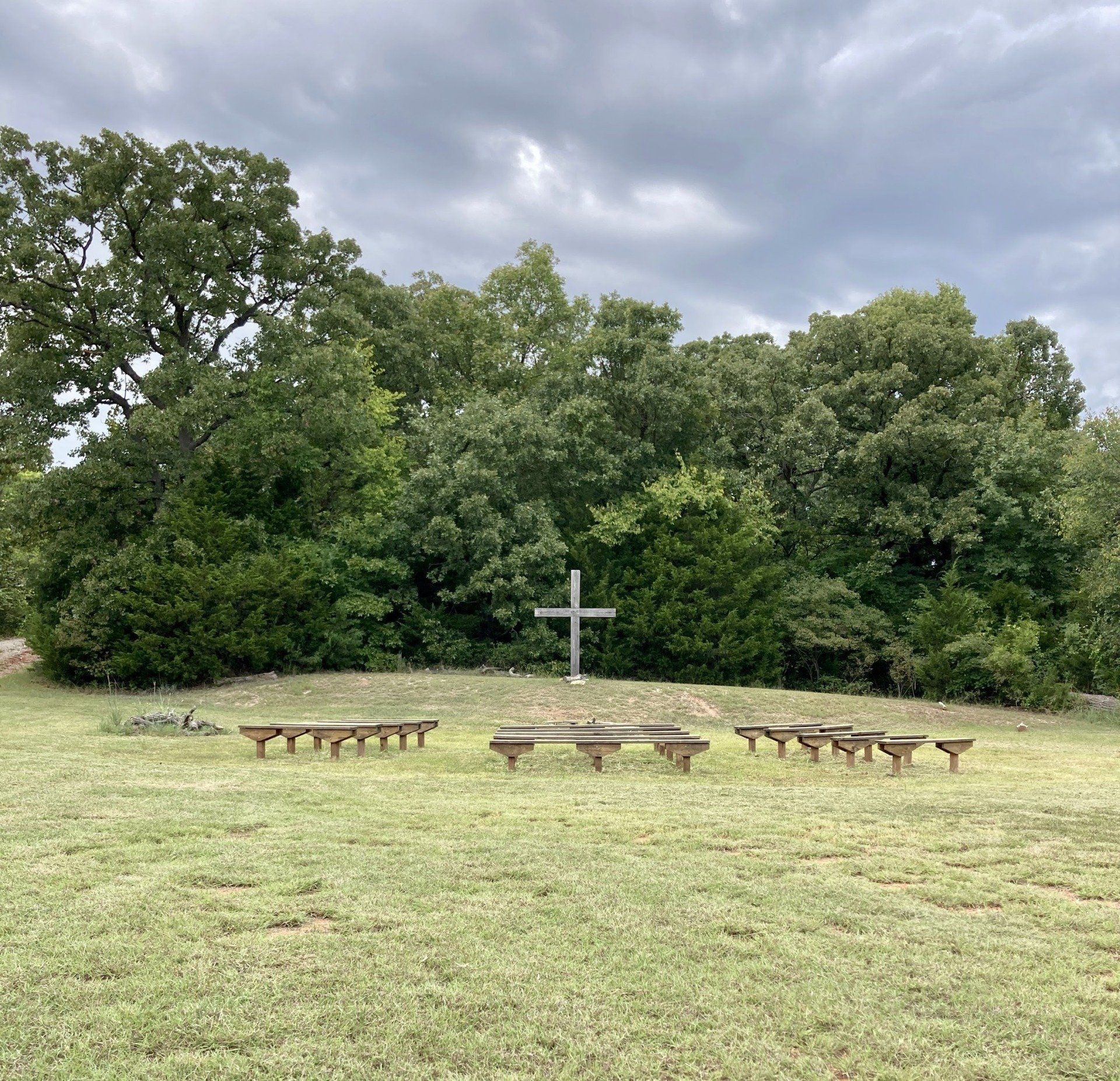 A cross on a small hill surrounded by trees, with wooden benches in front. Overcast sky.