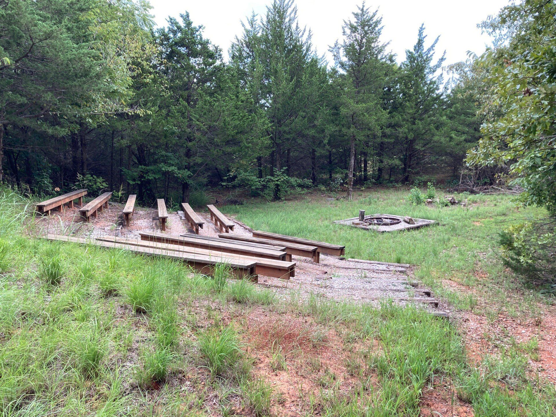 Outdoor amphitheater with wooden benches, fire pit, and trees in background.