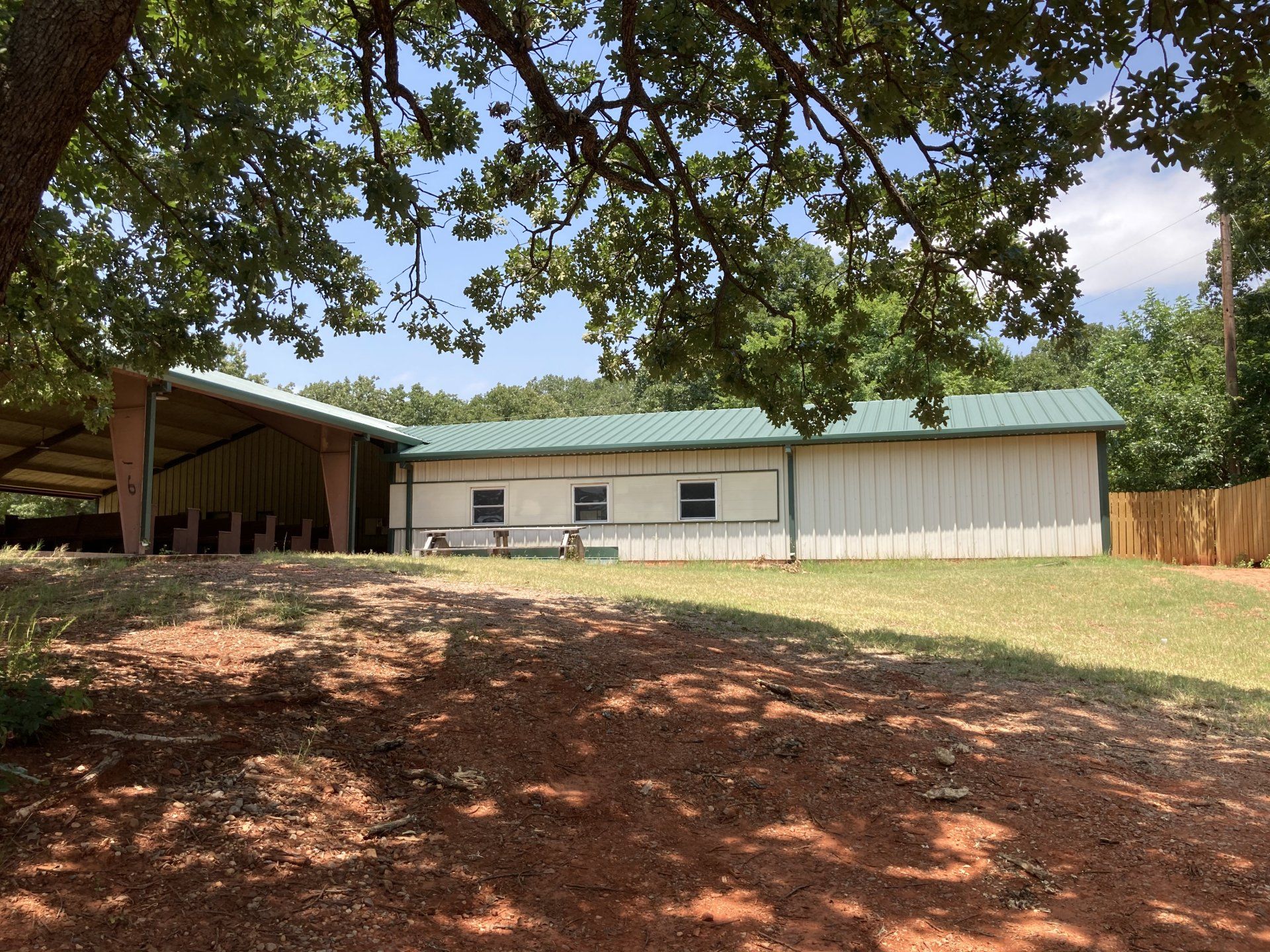 A one-story white building with a green roof, covered patio, and wooden fence, under tree shade.