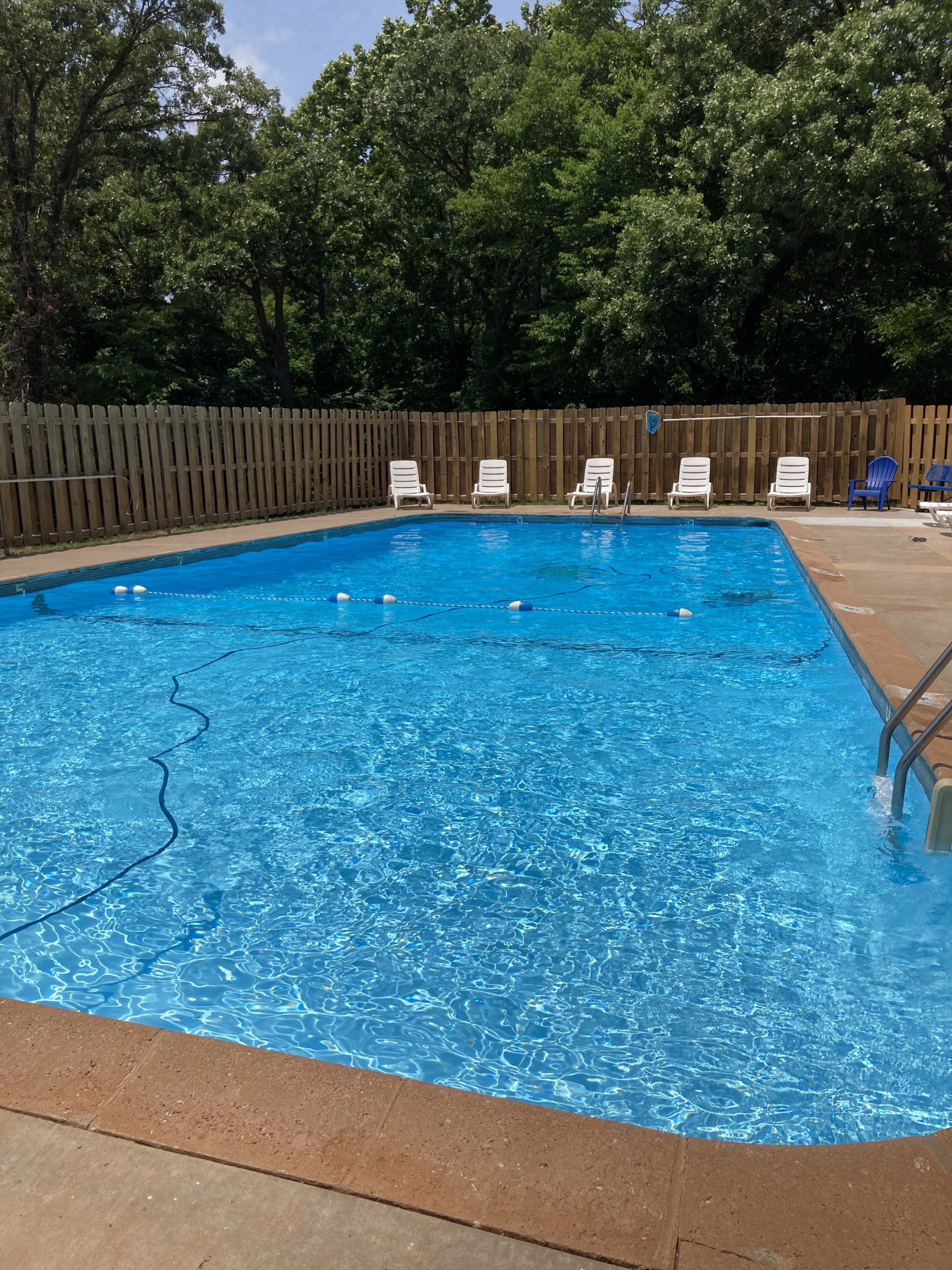 Swimming pool with blue water, surrounded by a wooden fence and trees, with empty chairs.