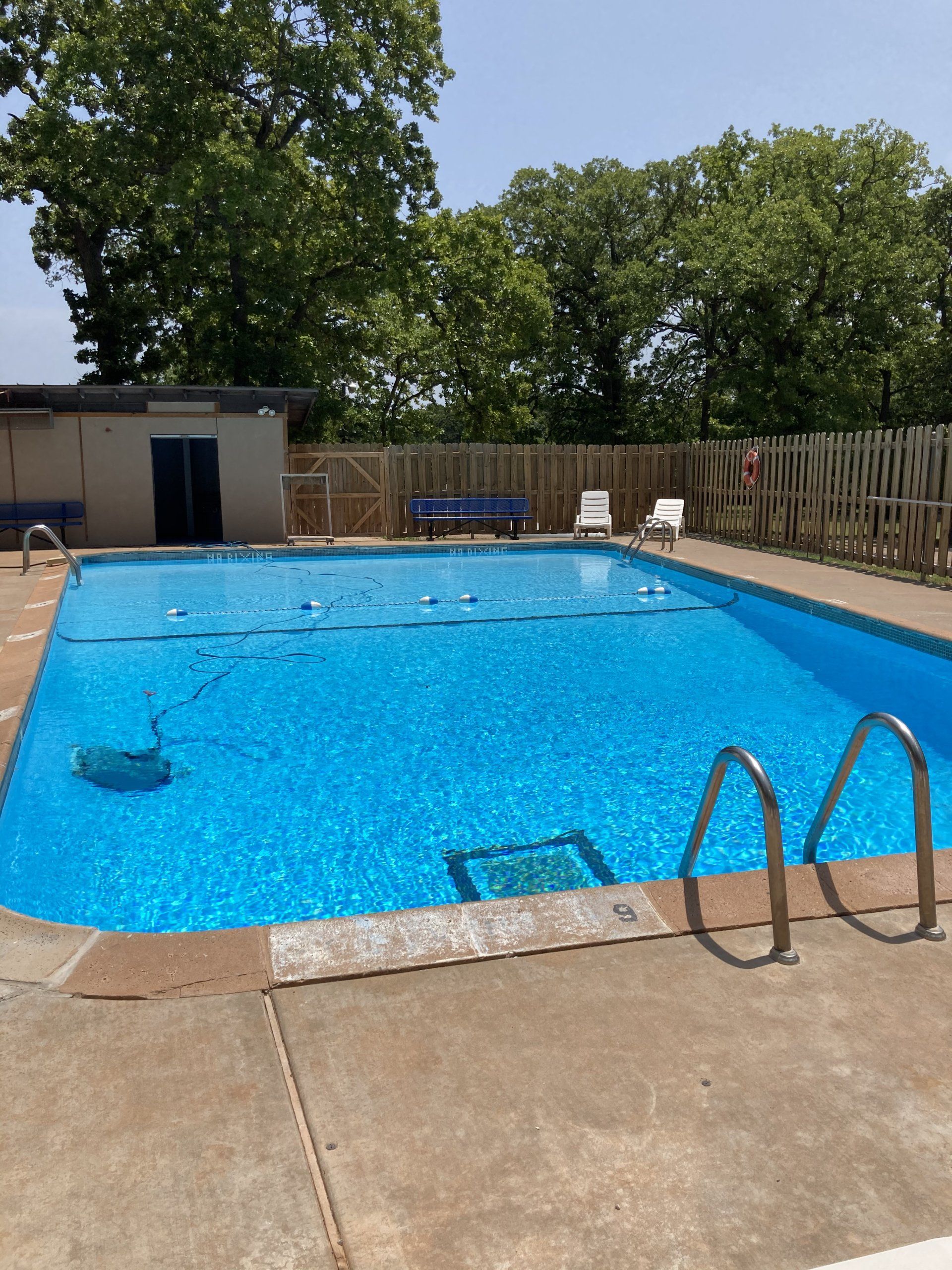 Swimming pool with blue water and surrounding concrete, fence, and trees under a clear sky.
