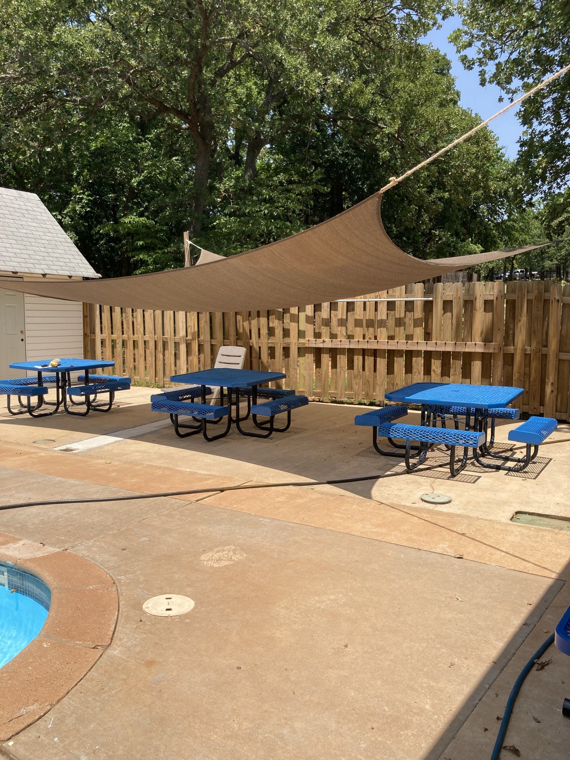 Outdoor dining area with blue picnic tables, a shade sail, and a wooden fence.