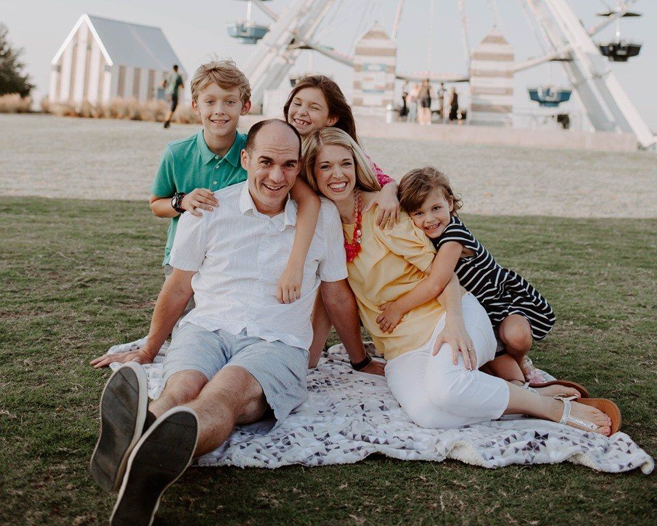 Family of five smiles together on a blanket outside.
