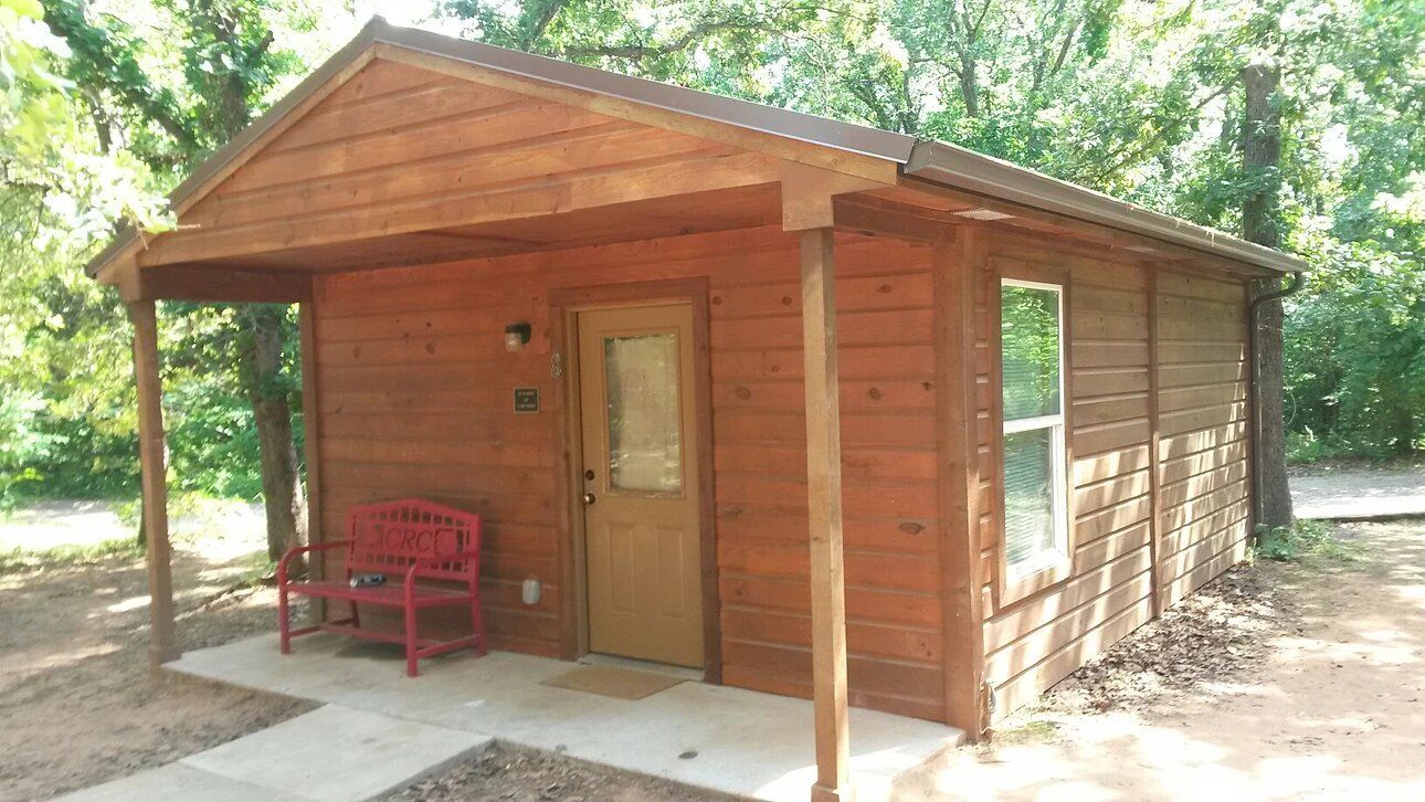 Small brown cabin with a porch and red bench; set in a wooded area.