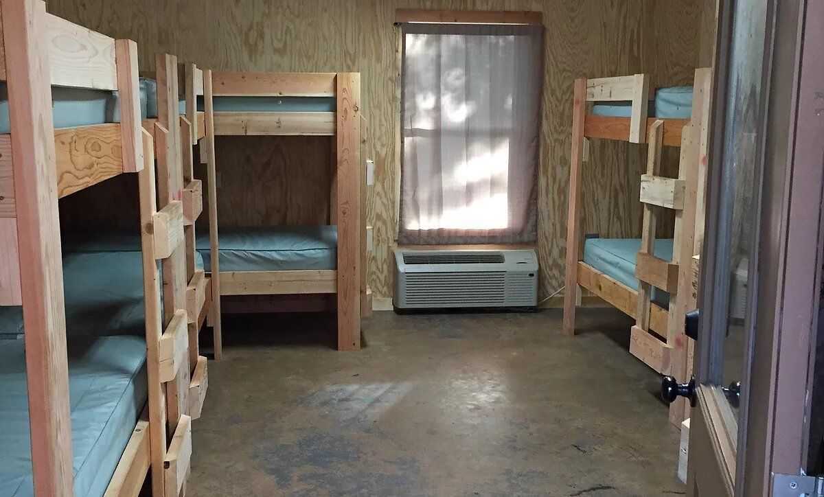 Wooden bunk beds in a room with a window, air conditioner, and plain walls and floor.