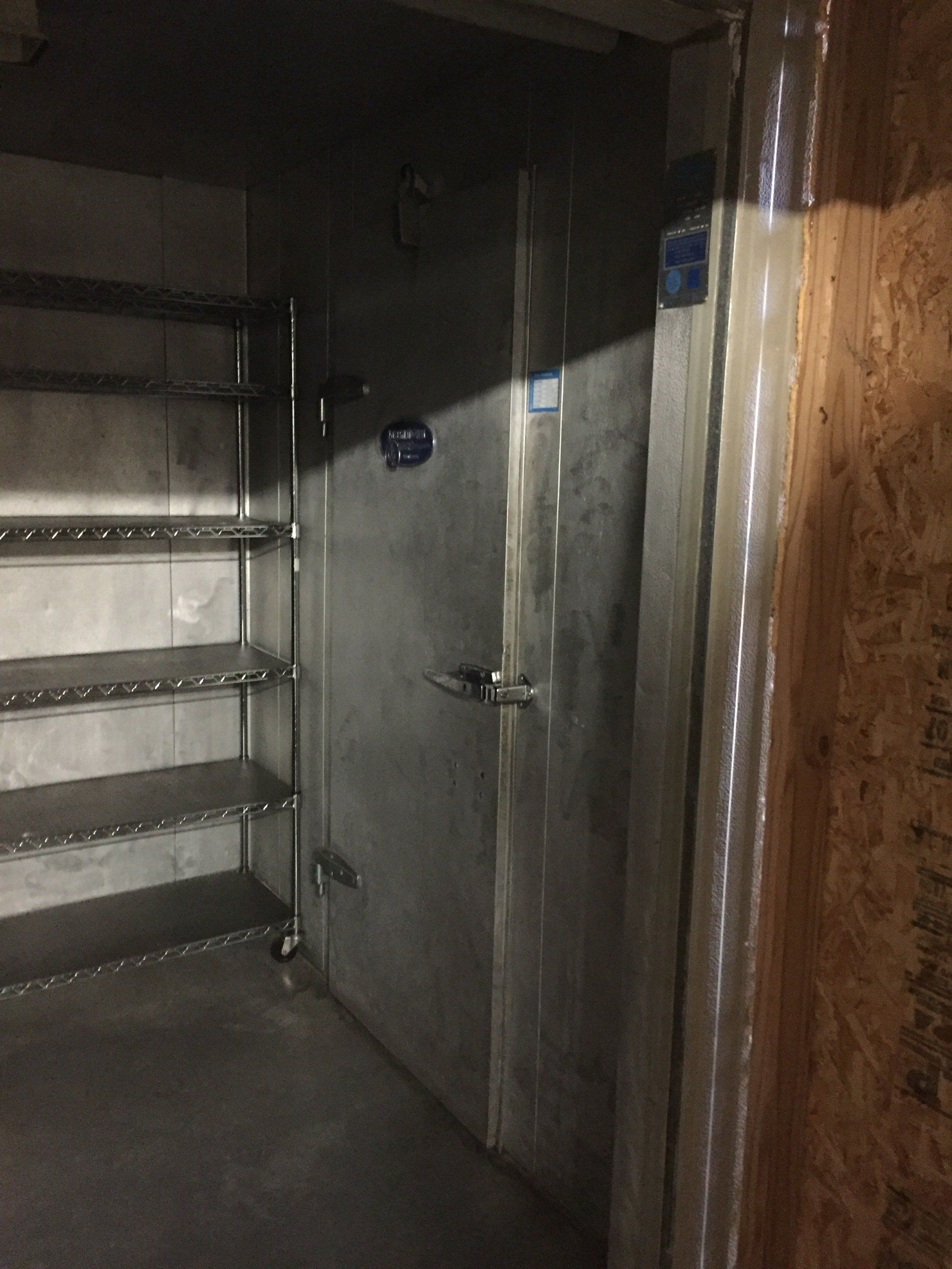 Walk-in cooler with shelves on the left, metal door in the center, and a partially visible wooden wall on the right.