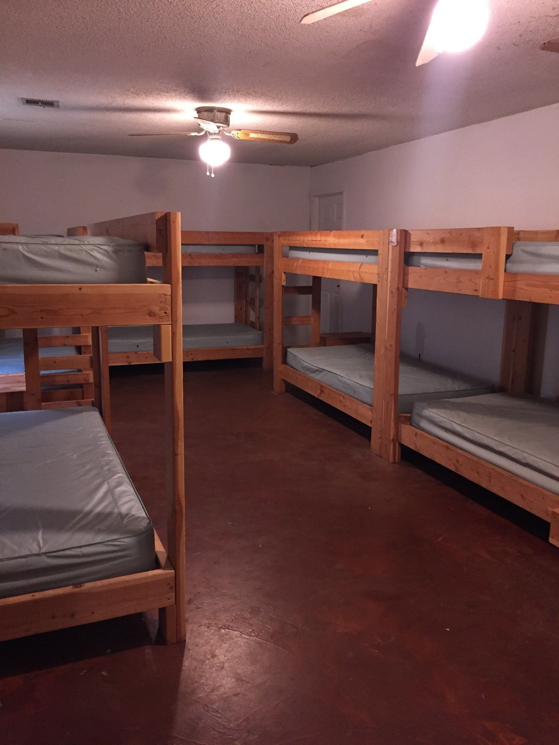 Dorm room with several wooden bunk beds, teal mattresses, and a ceiling fan. Brown floor, white walls.
