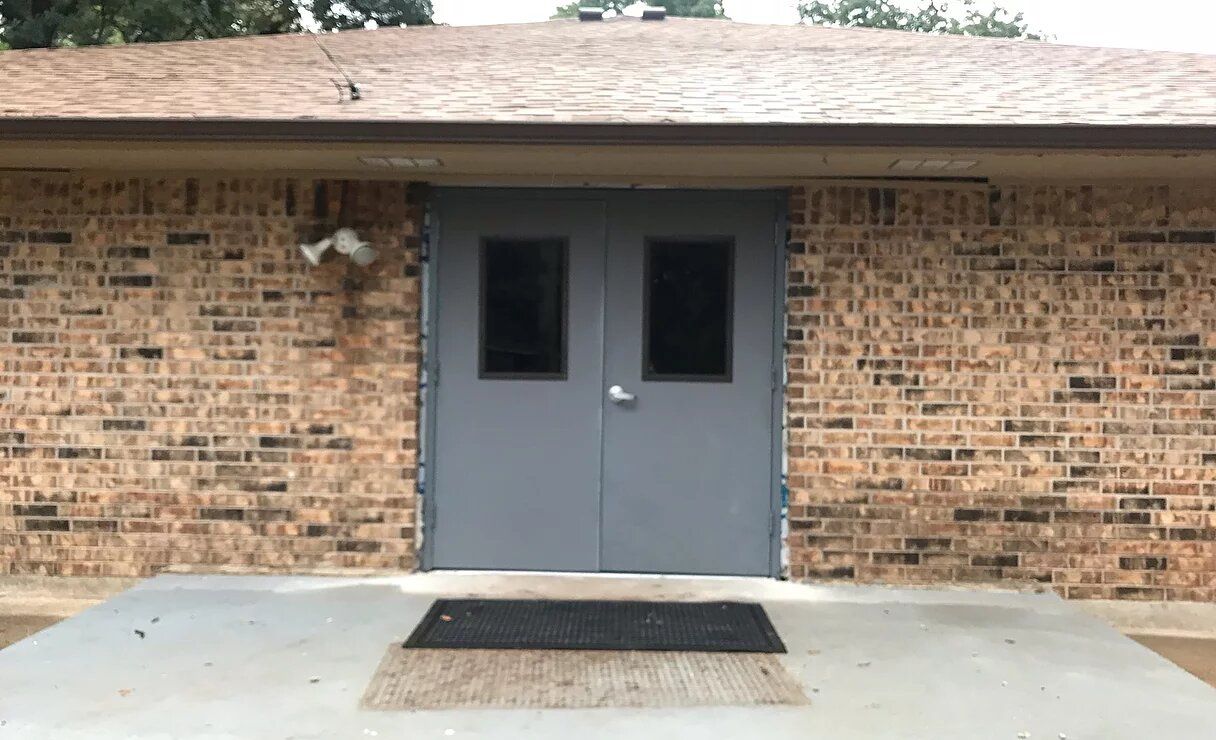 Gray double doors with small windows on a brick building, under a brown roof. A black doormat is in front.