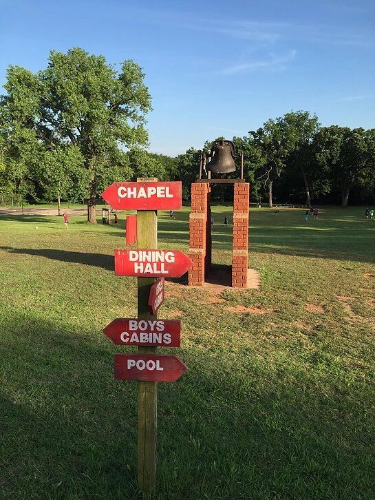 Signpost with red arrows pointing towards chapel, dining hall, boys cabins, and pool. A bell is mounted on brick pillars.