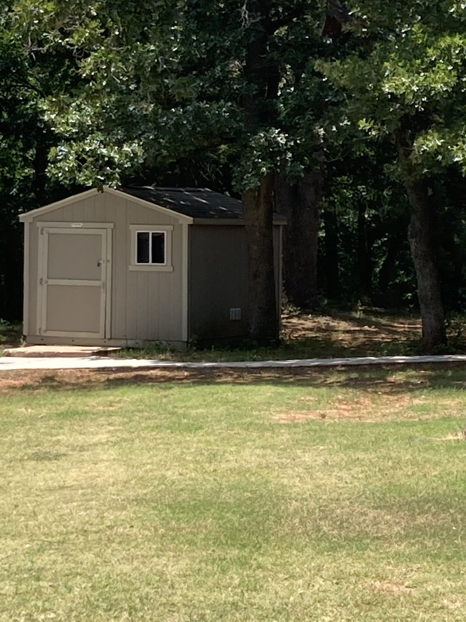 Small beige shed with white door and window, set on grassy ground near trees.