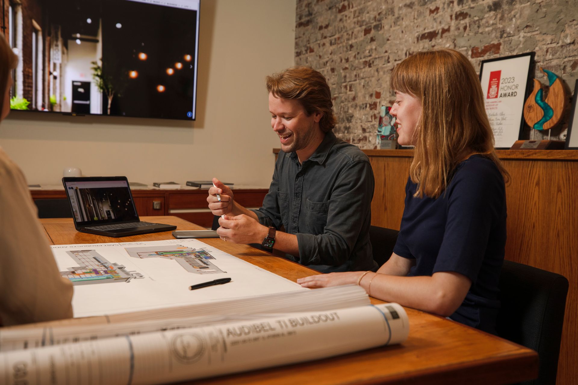 Three people at a table reviewing architectural plans. Man laughs, woman smiles, computer and TV in background.