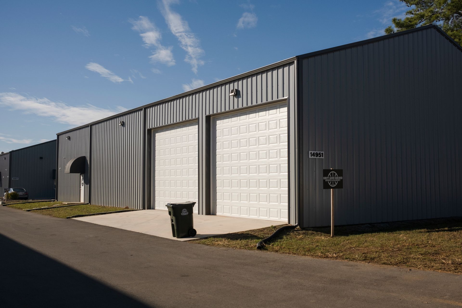 Gray metal warehouse building with two garage doors, a trash bin, and a sign on a sunny day.