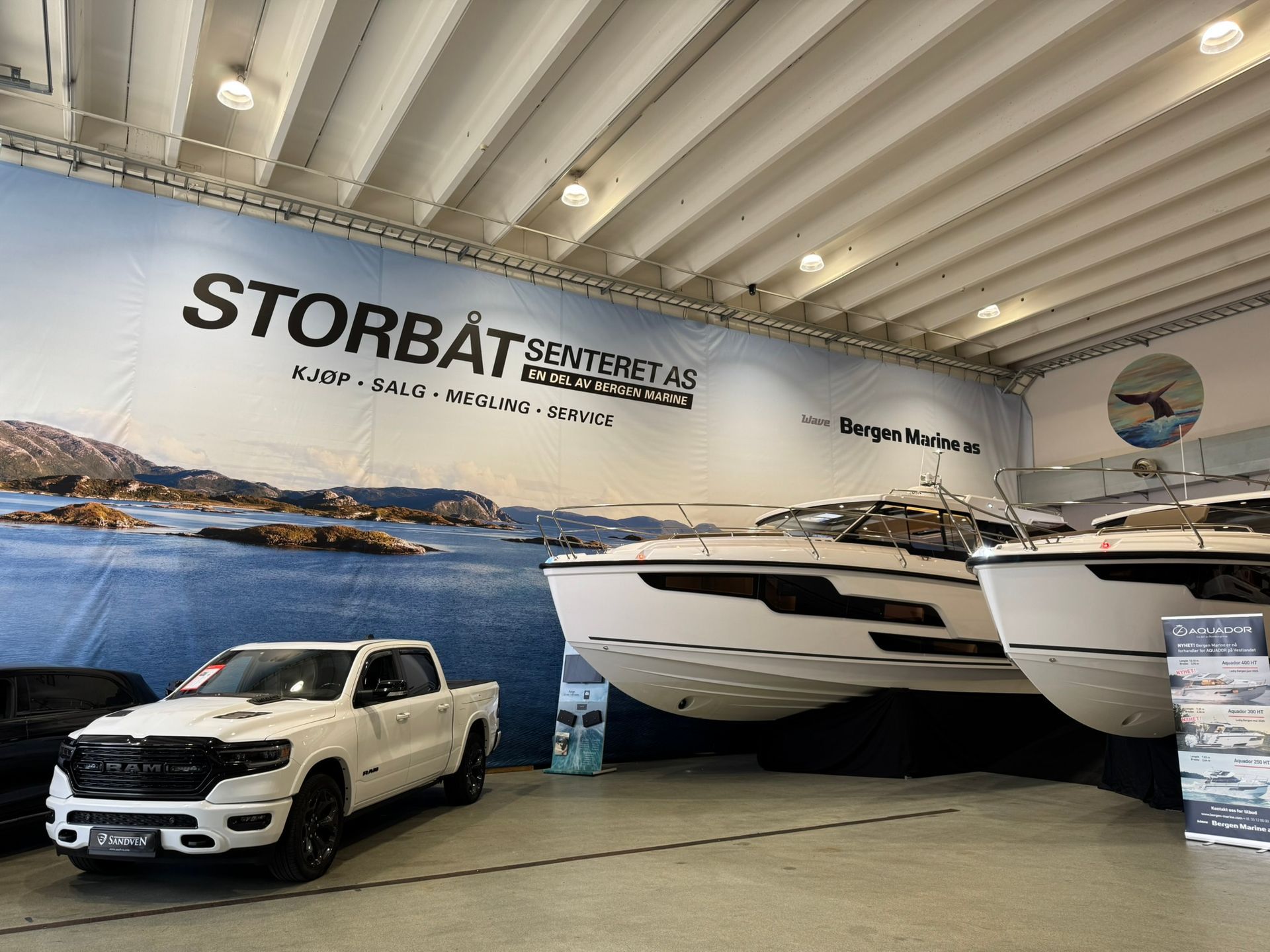En hvit lastebil og to båter utstilt inni, med vann og fjell som bakgrunn.