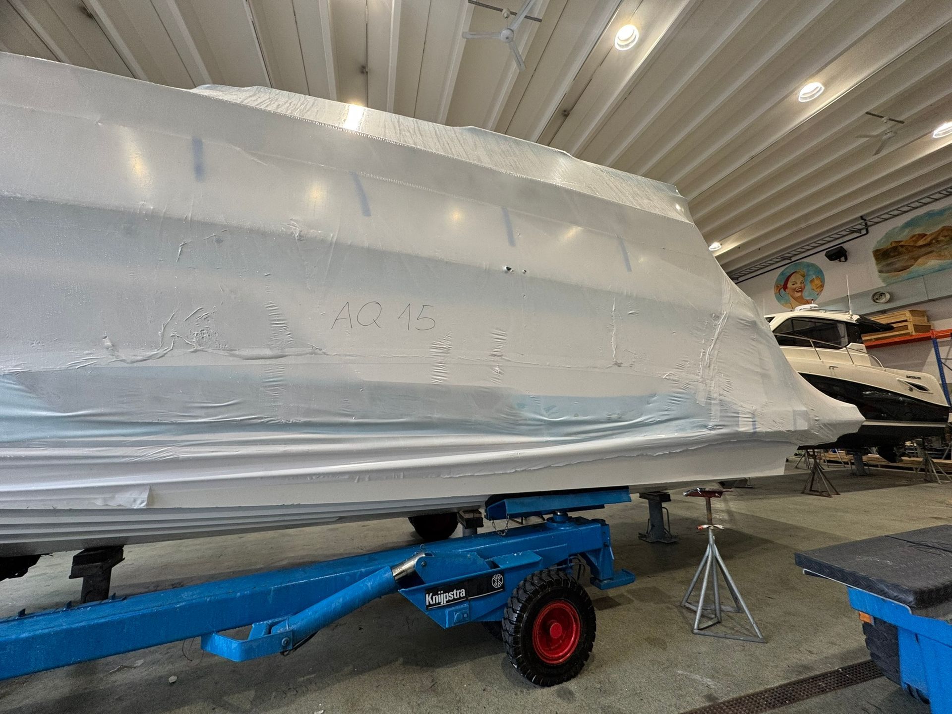 Båt pakket inn i hvit krympeplast på en blå tilhenger, inne i en stor hvit bygning.