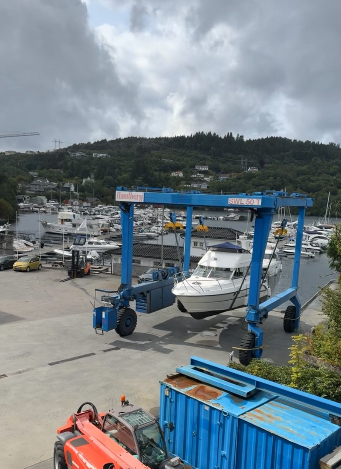 En blå båtheis som løfter en hvit båt i en havn, med grønne åser i bakgrunnen.