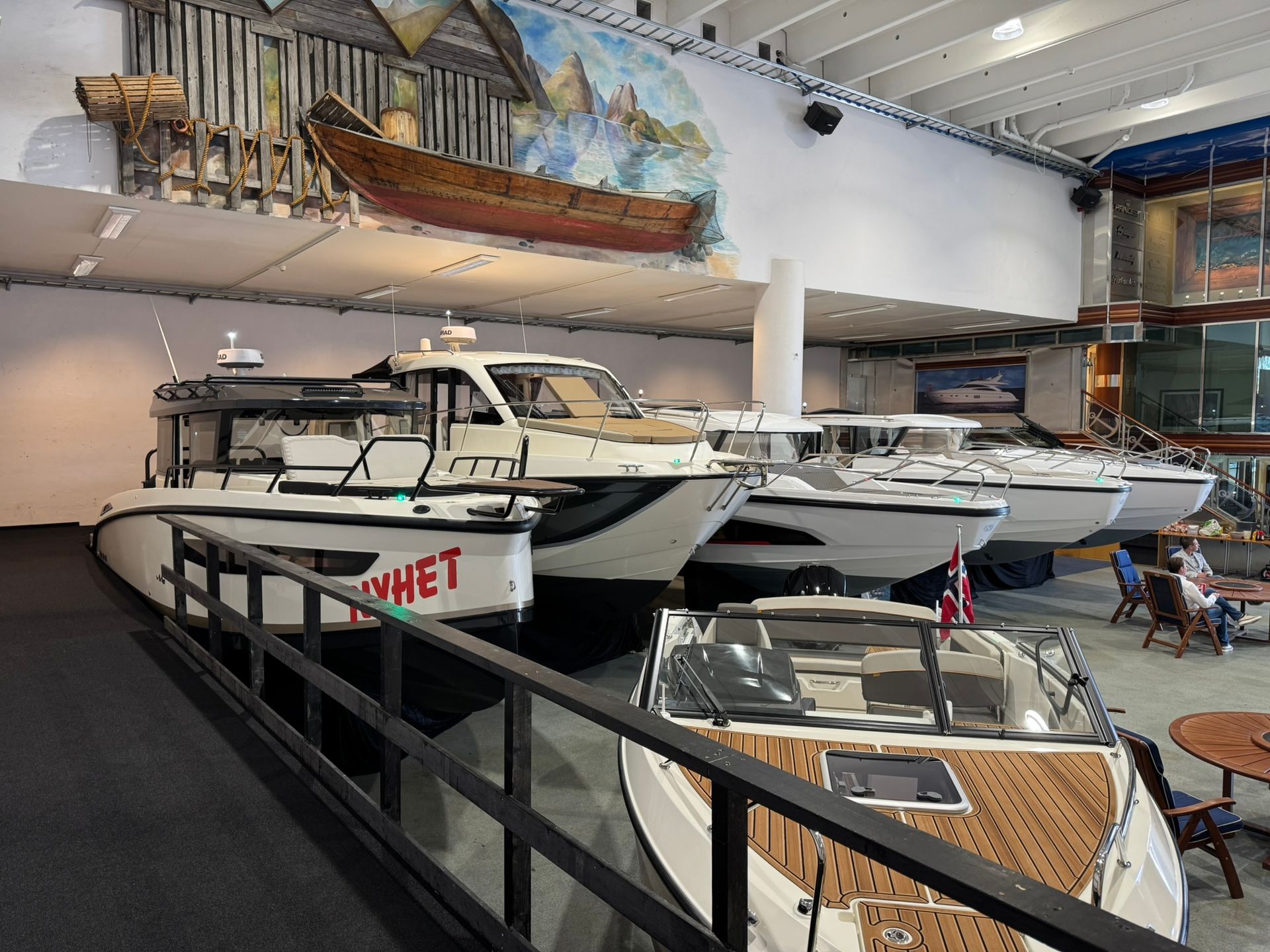 Båter utstilt innendørs, noen hvite, én med rød skrift, under et veggmaleri og en hengende trebåt.