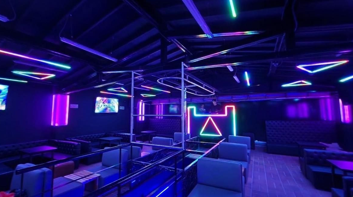 Dentro de un club con luces de neón. Colores azul y morado iluminan el espacio. Asientos vacíos y un poste central.