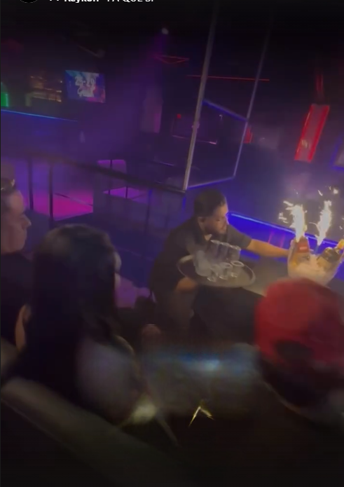 Persona vestida de forma oscura que se acerca a una mesa con bengalas en un club con luces de colores.