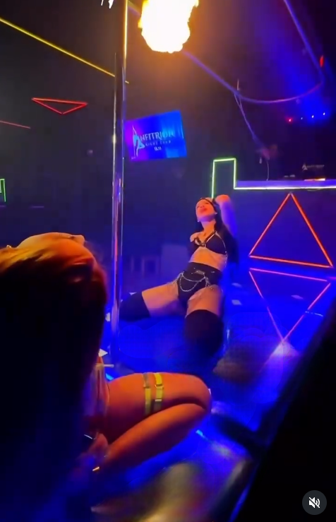 Una mujer baila en un tubo en una discoteca con luces de colores. Otra mujer observa desde abajo.