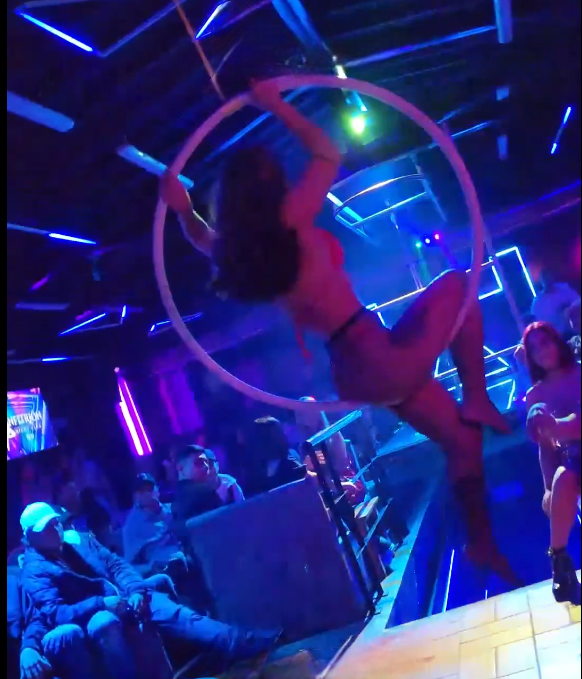 Mujer realizando una danza aérea con aro en un club poco iluminado con un espectáculo de luces azules y violetas.