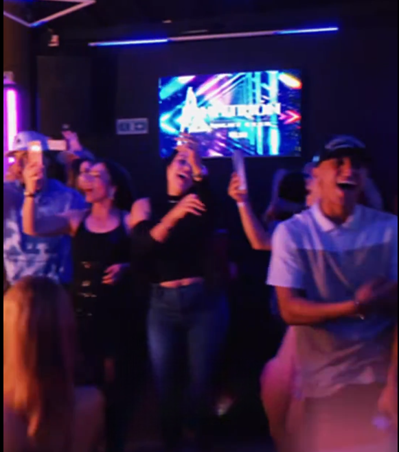 Gente cantando y sosteniendo teléfonos en un karaoke. Luces de neón, una pantalla con texto y un ambiente oscuro.