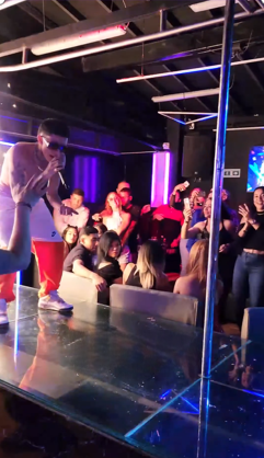 Hombre actuando en el escenario de un club, con el público vitoreando. Luces brillantes, fondo oscuro.