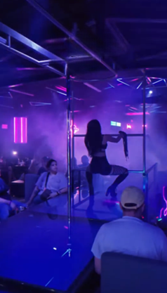 Una persona actúa en un poste en un club con iluminación azul y violeta y público.