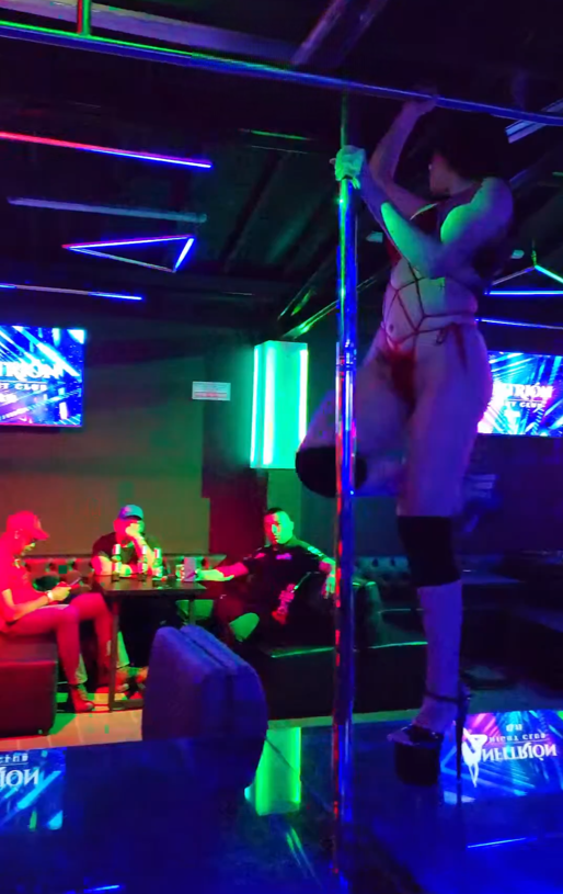 Una persona realiza pole dance en una discoteca con luces de neón; varias personas observan.