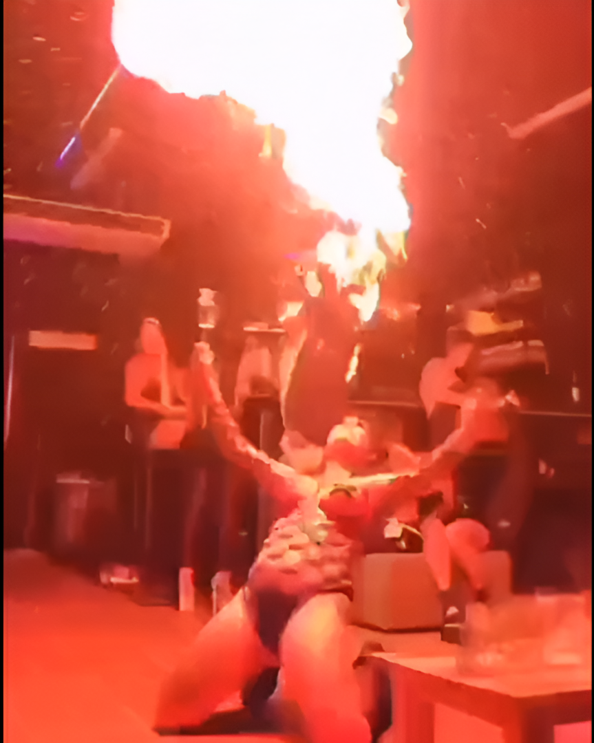 Mujer realizando un acto de escupir fuego en un club poco iluminado; llamas brotan de su boca.