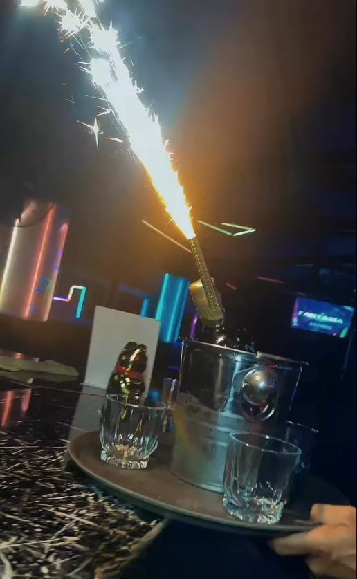 Botella con bengala en un cubo sobre una bandeja, servida con vasos en un club oscuro.