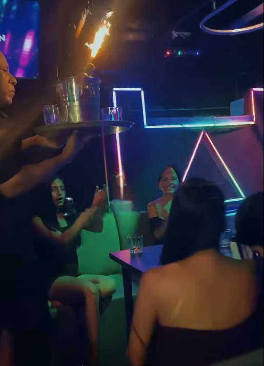 Camarero llevando una botella en llamas en una bandeja en un club nocturno poco iluminado con luces de neón.