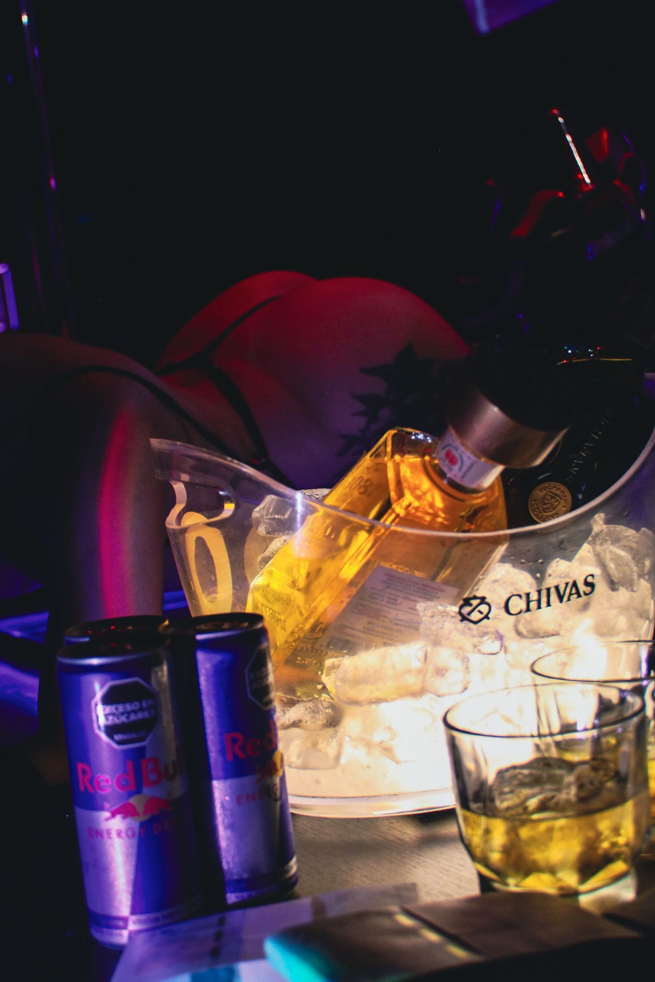 Una botella de whisky en un cubo de hielo, latas de Red Bull y una bebida en un vaso en una barra oscura con una persona de fondo.