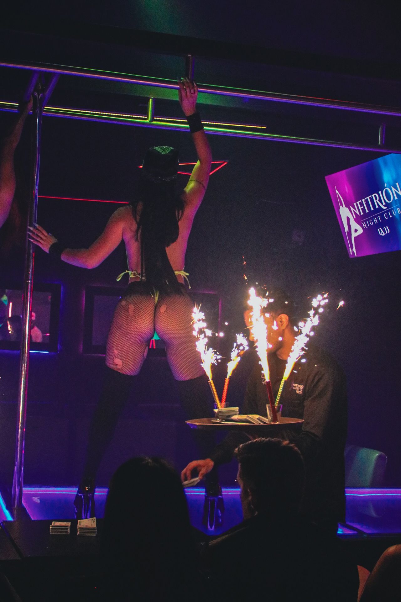 Una persona baila en barra en un club con poca luz. Hay bengalas en una bandeja sostenida por alguien desde abajo.