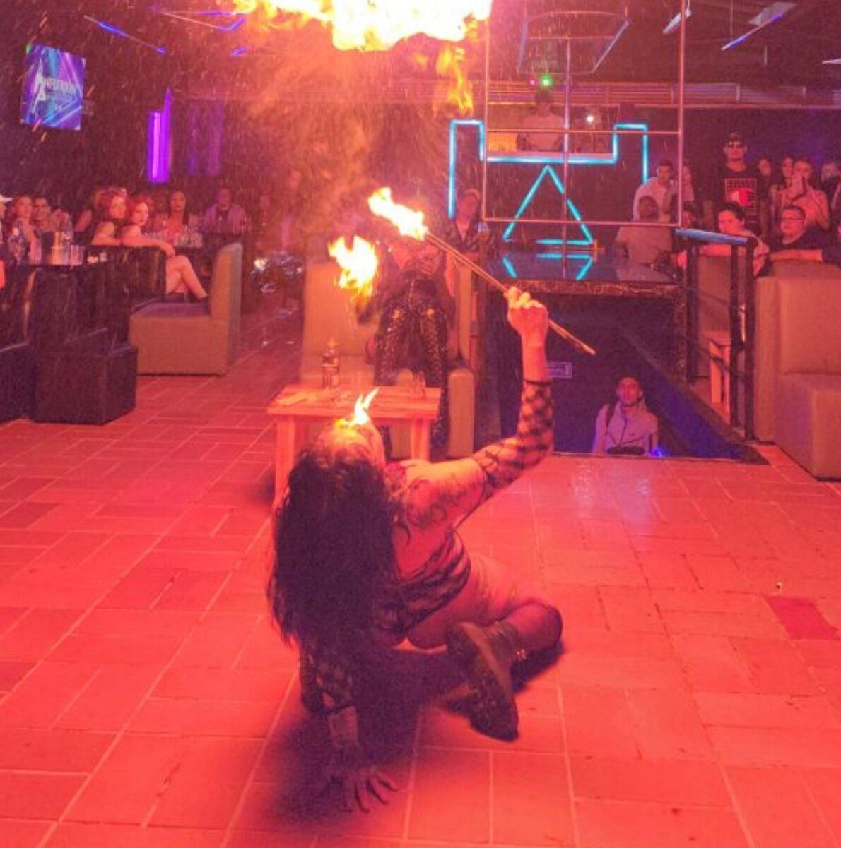 Un artista se arrodilla, lanzando fuego en un club iluminado con luz roja. Llamas brotan de su boca. El público observa.