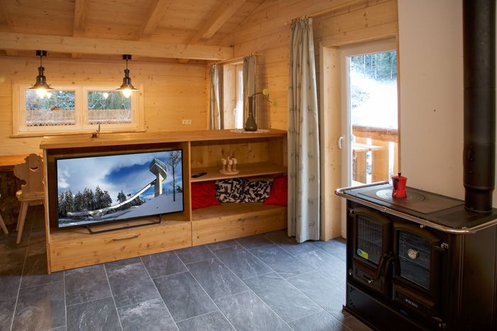Ein gemütliches, holzgetäfeltes Zimmer mit Fernseher, Holzregalen und einem schwarzen Holzofen auf grauem Fliesenboden.